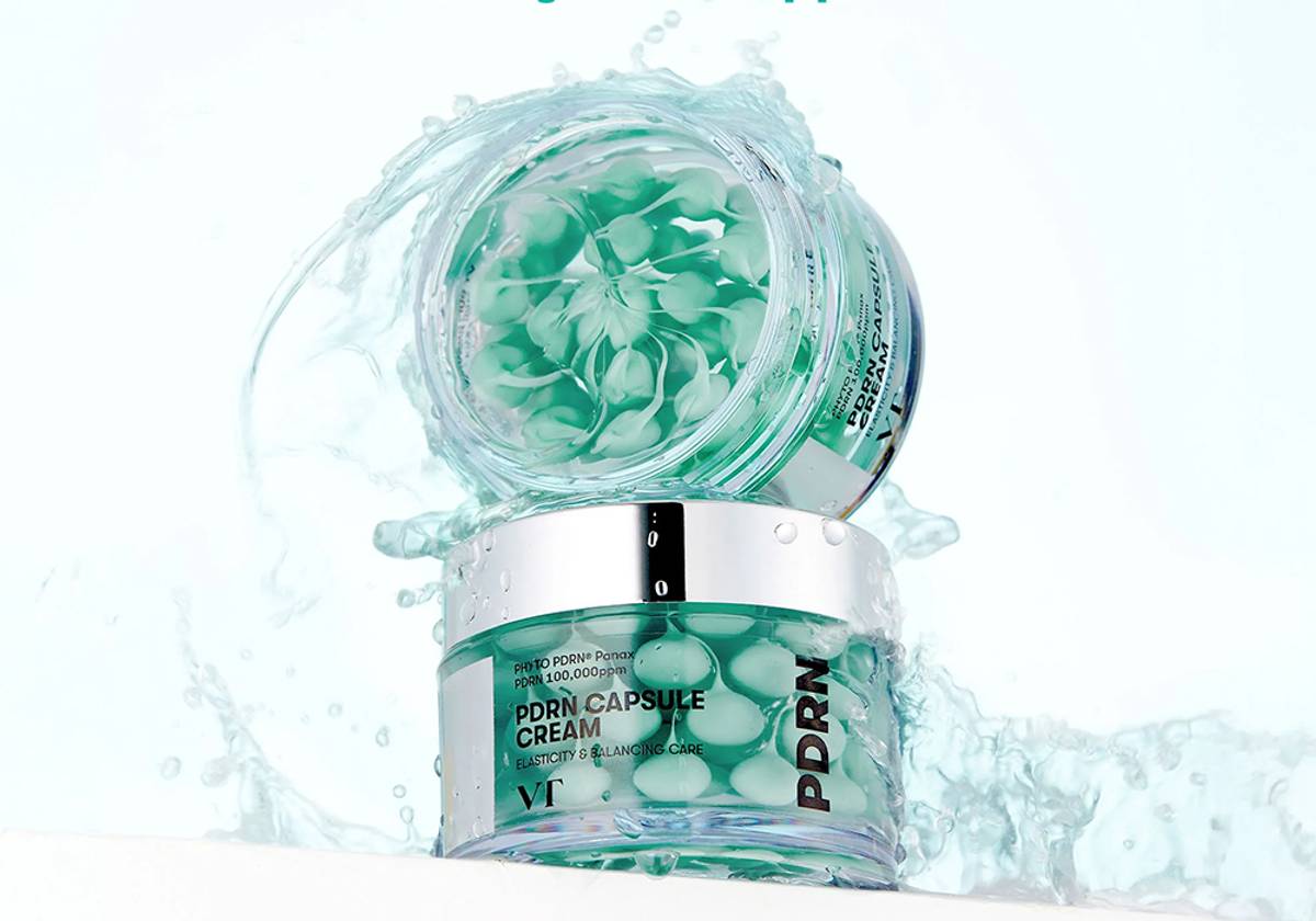 An open jar of VT Cosmetics PDRN Capsule Cream showing green gel capsules inside, surrounded by water splashes against a light blue background.