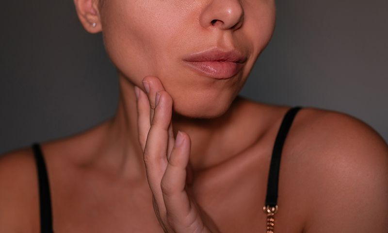 Young woman touching her cheek, showing defined jawline and buccal area on a dark grey background. Concept of buccal fat removal, face contouring and aesthetic facial procedures