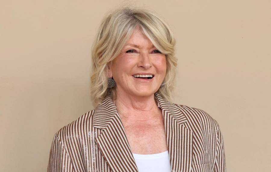 Martha Stewart smiling against a beige backdrop, wearing a brown-and-white striped blazer over a white top with beaded drop earrings.