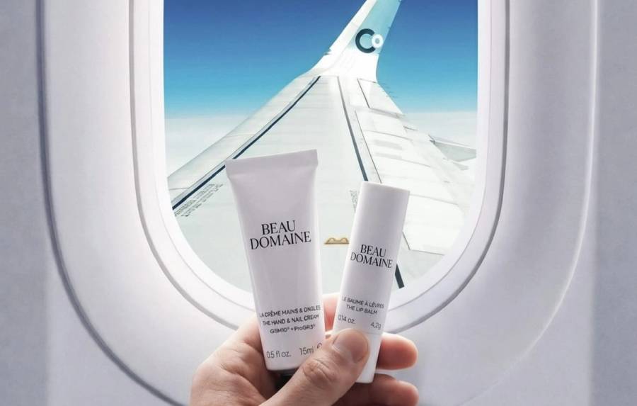 A hand holds Beau Domaine hand cream and lip balm in front of an airplane window, with a La Compagnie wing visible against a bright blue sky