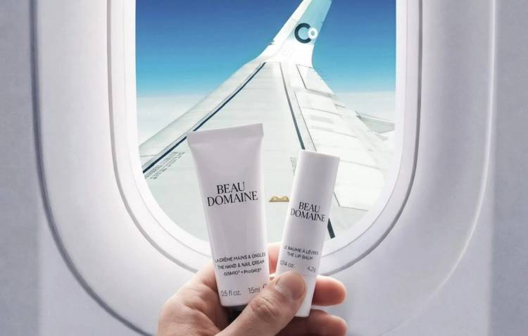 A hand holds Beau Domaine hand cream and lip balm in front of an airplane window, with a La Compagnie wing visible against a bright blue sky