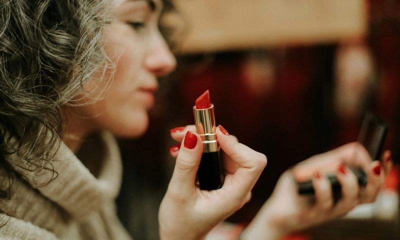 Woman with gray-ish hair and red nails holding red lipstick and looking in a makeup compact