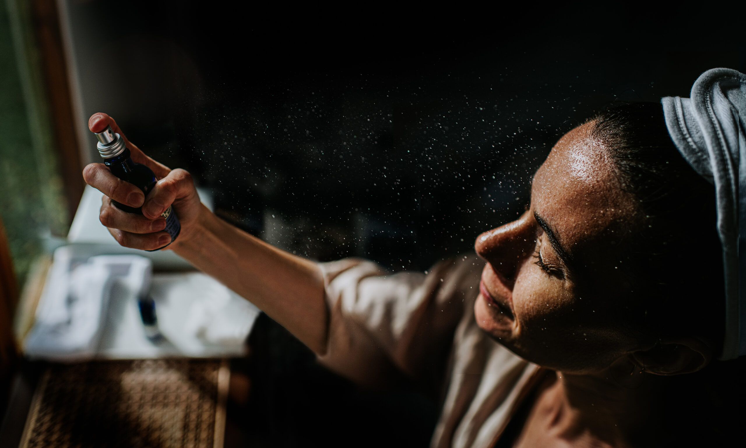 Moody, cinematic image of a woman holding a spray bottle, and spritzing moisture on to her face. Shadows provide a space for copy.