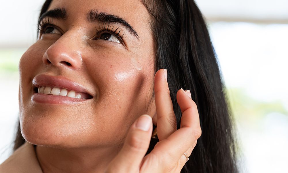 Happy woman applying moisturizing cream on her face, enjoying a moment of self-care and enhancing her skin's natural beauty indoors