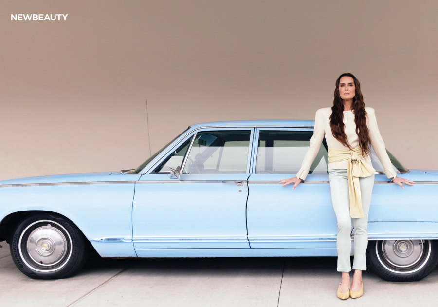 Brooke Shields poses leaning against a vintage baby blue car wearing a white blazer, cream top and light jeans for NewBeauty's summer 2026 cover shoot