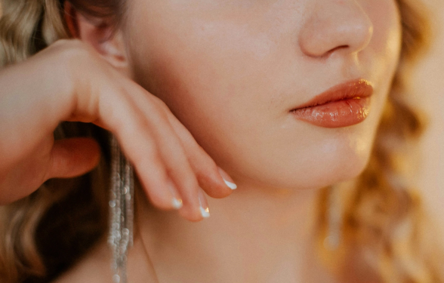 Close-up of a woman resting her hand beneath her chin, wearing glossy nude lipstick and dangling silver earrings in warm golden light.
