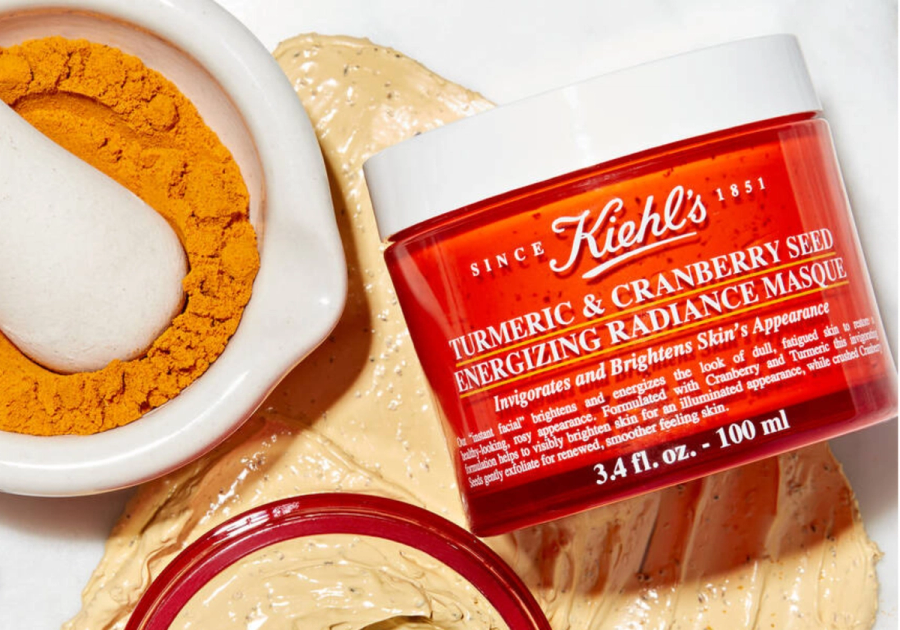 Kiehl's Turmeric & Cranberry Seed Energizing Radiance Masque jar surrounded by turmeric powder and product texture swatches on a white background.