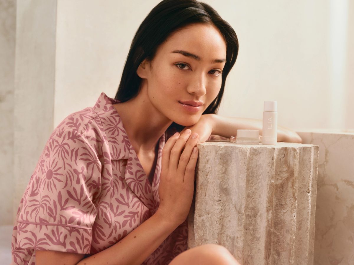 Model in Eberjey x ILIA pink silk pajama set applying ILIA skin care in a softly lit bedroom setting.