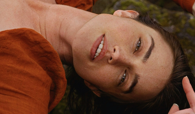 Close-up of a woman with slicked-back dark hair lying outdoors in a rust-colored top and looking at the camera.