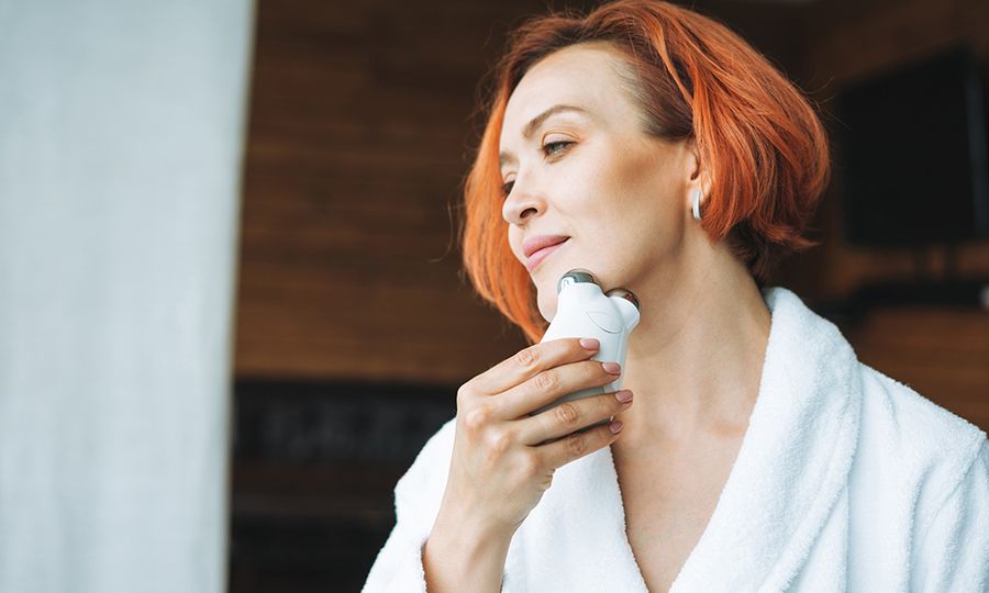 Beauty portrait of smiling woman 35 year in white bathrobe with clean fresh face and hands with red hair doing fasial massage with microcurrent facial massager at the bath room, treat yourself, home body care