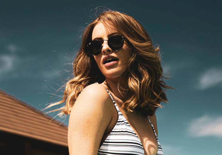 Woman with softly highlighted brond hair and loose waves wearing sunglasses outdoors under a bright summer sky, showcasing a dimensional, sun-kissed hair color.