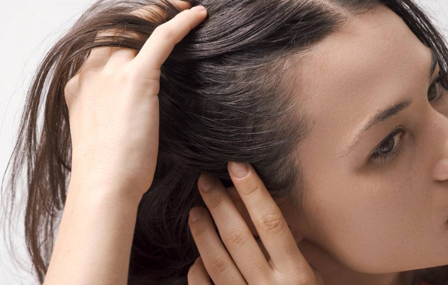 Close-up of a woman parting her dark hair with her fingers to examine her scalp.