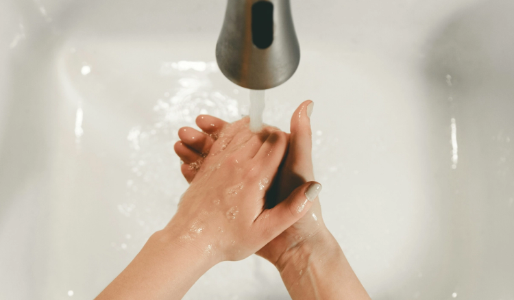 Overhead view of two hands being washed under running water in a white sink, fingers laced together with soap suds visible.