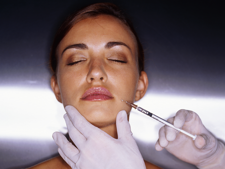 A woman receiving a dermal filler injection near the mouth area, administered by a gloved practitioner, eyes closed against a dark background.