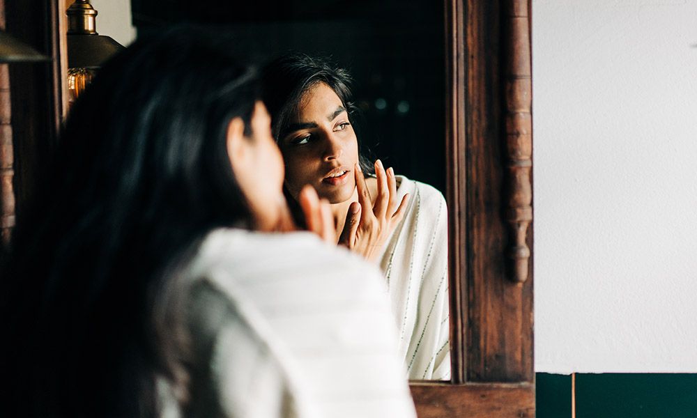 Young woman in bathrobe applying moisturizing cream on face in the bathroom.