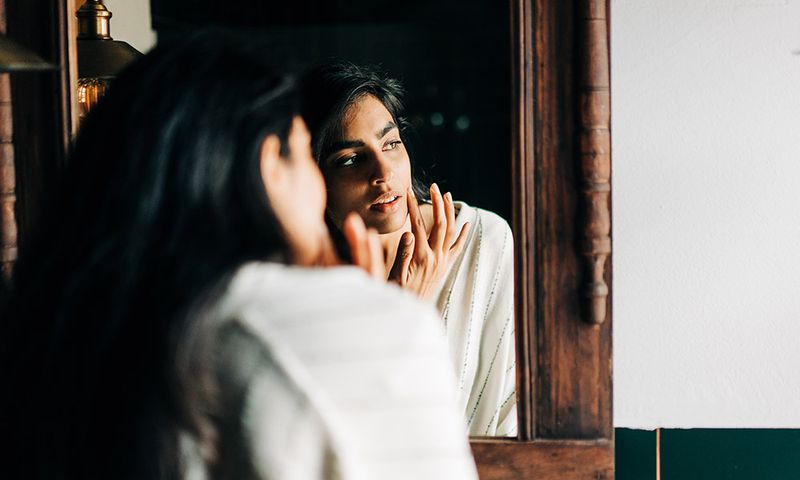 Young woman in bathrobe applying moisturizing cream on face in the bathroom.