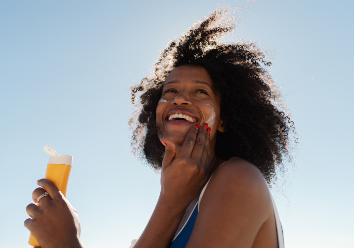 A smiling woman with curly hair applies sunscreen to her face outdoors against a blue sky, holding a yellow sunscreen bottle.