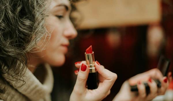 Woman with curly gray-brown hair holding an open red lipstick up to her face while looking in a mirror