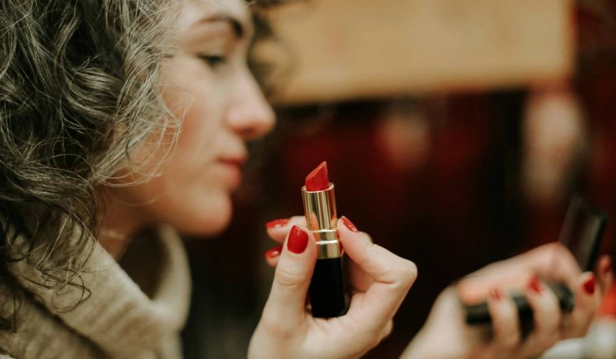 Woman with curly gray-brown hair holding an open red lipstick up to her face while looking in a mirror