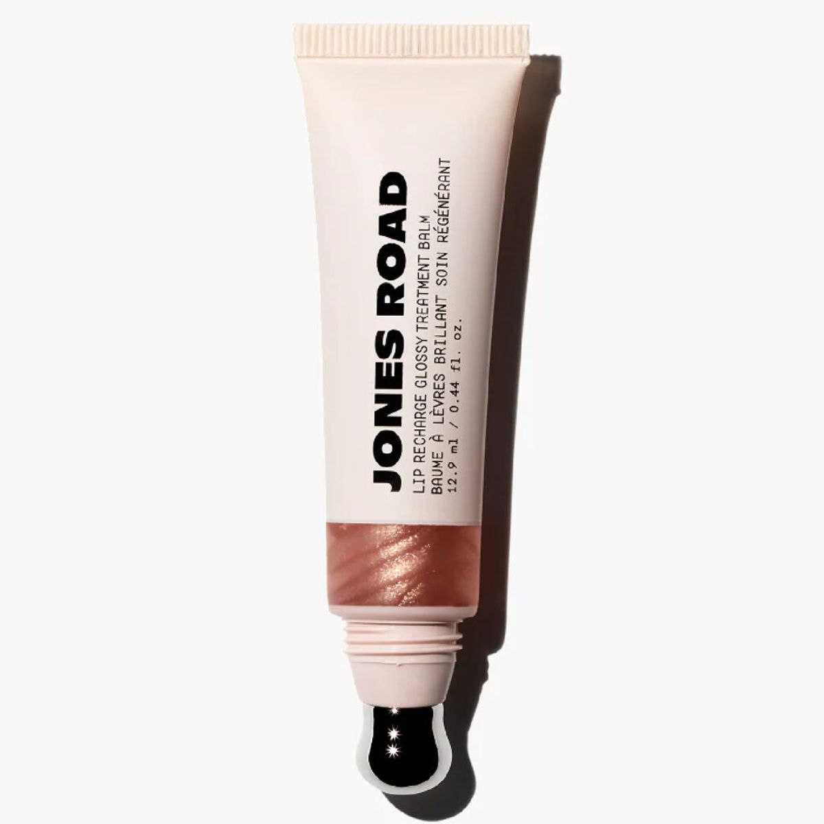 Jones Road Lip Recharge Glossy Treatment Balm in a beige tube with a translucent window showing a soft pink tint and a metal applicator tip, set against a light gray background.
