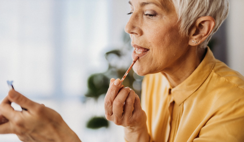 Mature woman applying lip color while looking in a mirror at home