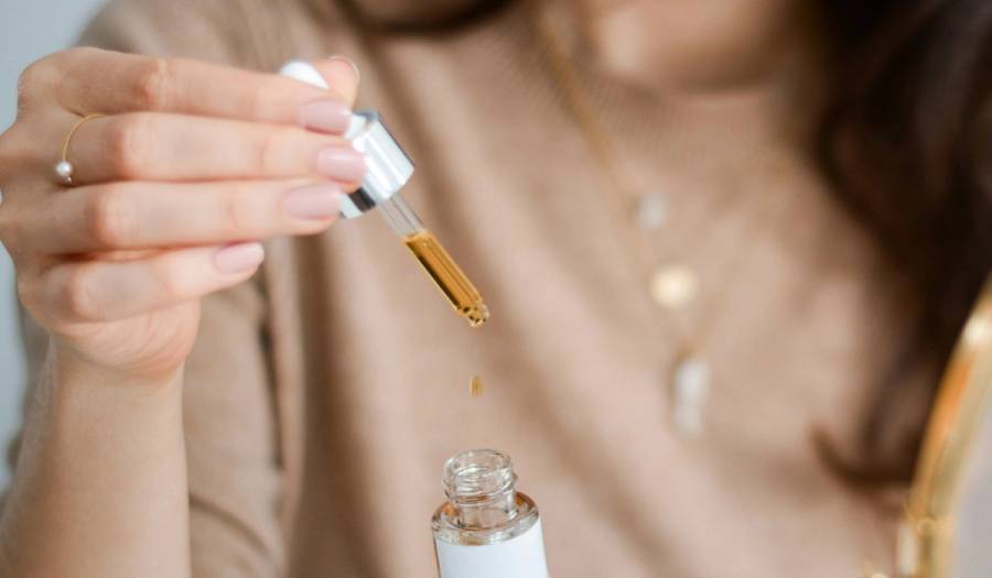 Close-up of a dropper releasing a drop of golden face serum into an open glass bottle, illustrating a fall hydration skin-care routine.