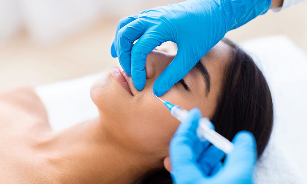 Nasolabial Fold Filler Procedure. Beautiful half-naked brunette indian woman gets cosmetic injection in nasolabial folds. Hyaluronic acid injection for facial rejuvenation treatment, closeup