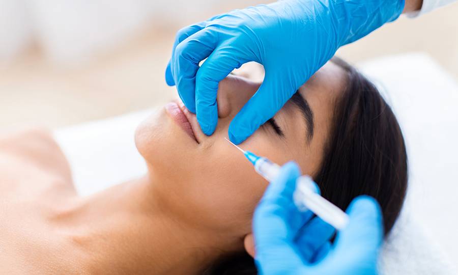 Nasolabial Fold Filler Procedure. Beautiful half-naked brunette indian woman gets cosmetic injection in nasolabial folds. Hyaluronic acid injection for facial rejuvenation treatment, closeup