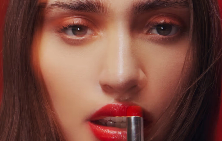 Close-up of a woman applying bright red lipstick against a deep red background.