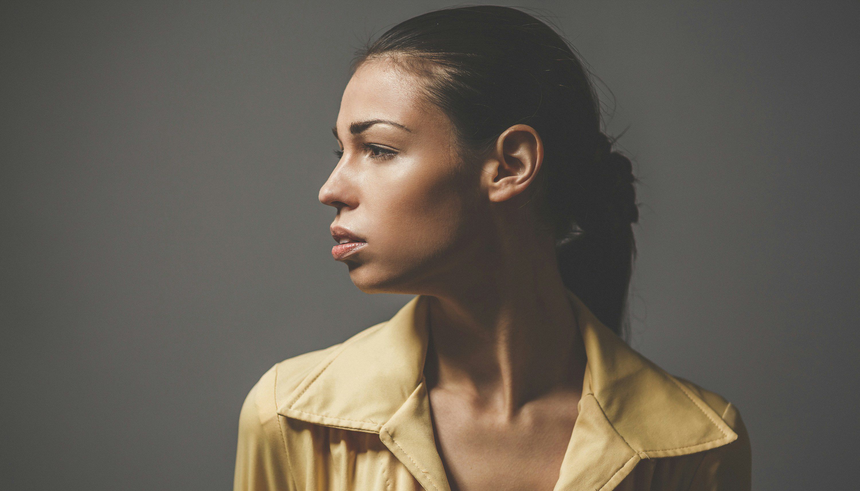 Side-profile portrait of a woman with her hair pulled back, wearing a yellow collared shirt and looking left against a gray background.