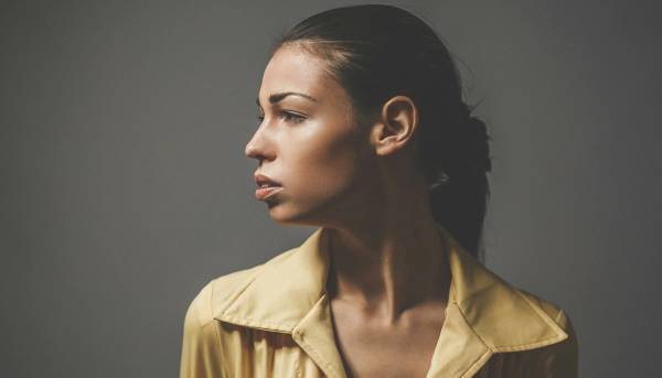 Side-profile portrait of a woman with her hair pulled back, wearing a yellow collared shirt and looking left against a gray background.