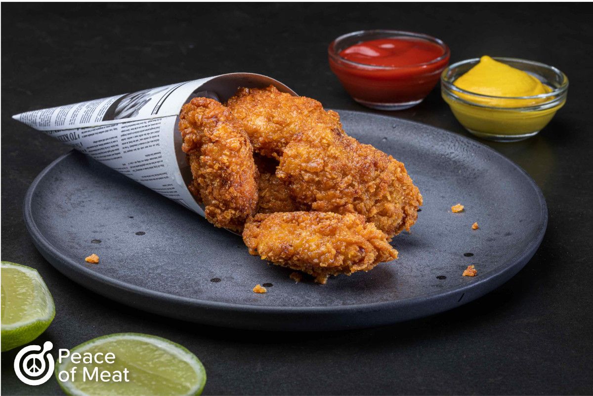 crispy plant-based chicken nuggets made from cultured chicken and plant-based protein. On plate in newspaper cone with ketchum, mustard, and lime on the side.