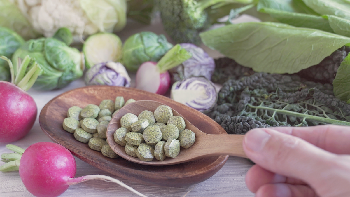 green pills on wooden spoon and bowl surrounded by cruciferous vegetables