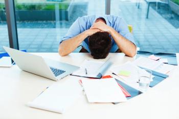 man with head on desk surrounded by papers and laptop indicating stress
