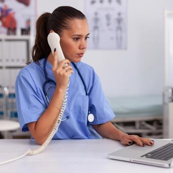 Image of a woman wearing scrubs sitting at a desk with a phone and laptop