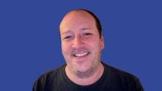 Image of a man wearing a black t-shirt smiling in front of a blue background