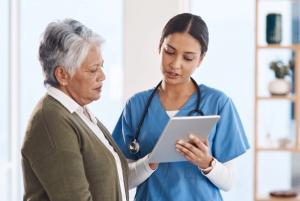 Image of a nurse and patient standing next to each other. The nurse is speaking and directing and directing the patient's attention toward a tablet.