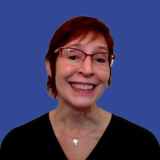Photo of a woman with red hair and glasses wearing a black blouse and cross necklace in front of a dark blue background
