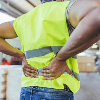 Photo of a man in a warehouse with a yellow vest holding his back in pain