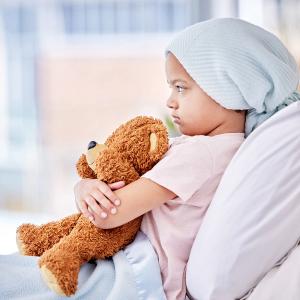Image of a pediatric patient with a headwrap with an upset face holding a teddy bear