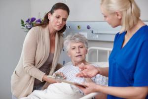 Photo of a nurse showing something on a clipboard to an older patient in bed and another woman, who is next to the patient and holding her hand.