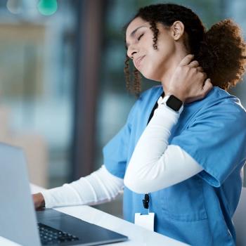 Photo of a woman in scrubs sitting in front of a laptop at a desk with her hand on her neck and an exhausted expression