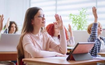 A child wearing glasses sits at a desk in a classroom, raising her hand. She is surrounded by her peers. Image credit: ©ihorvsn – stock.adobe.com