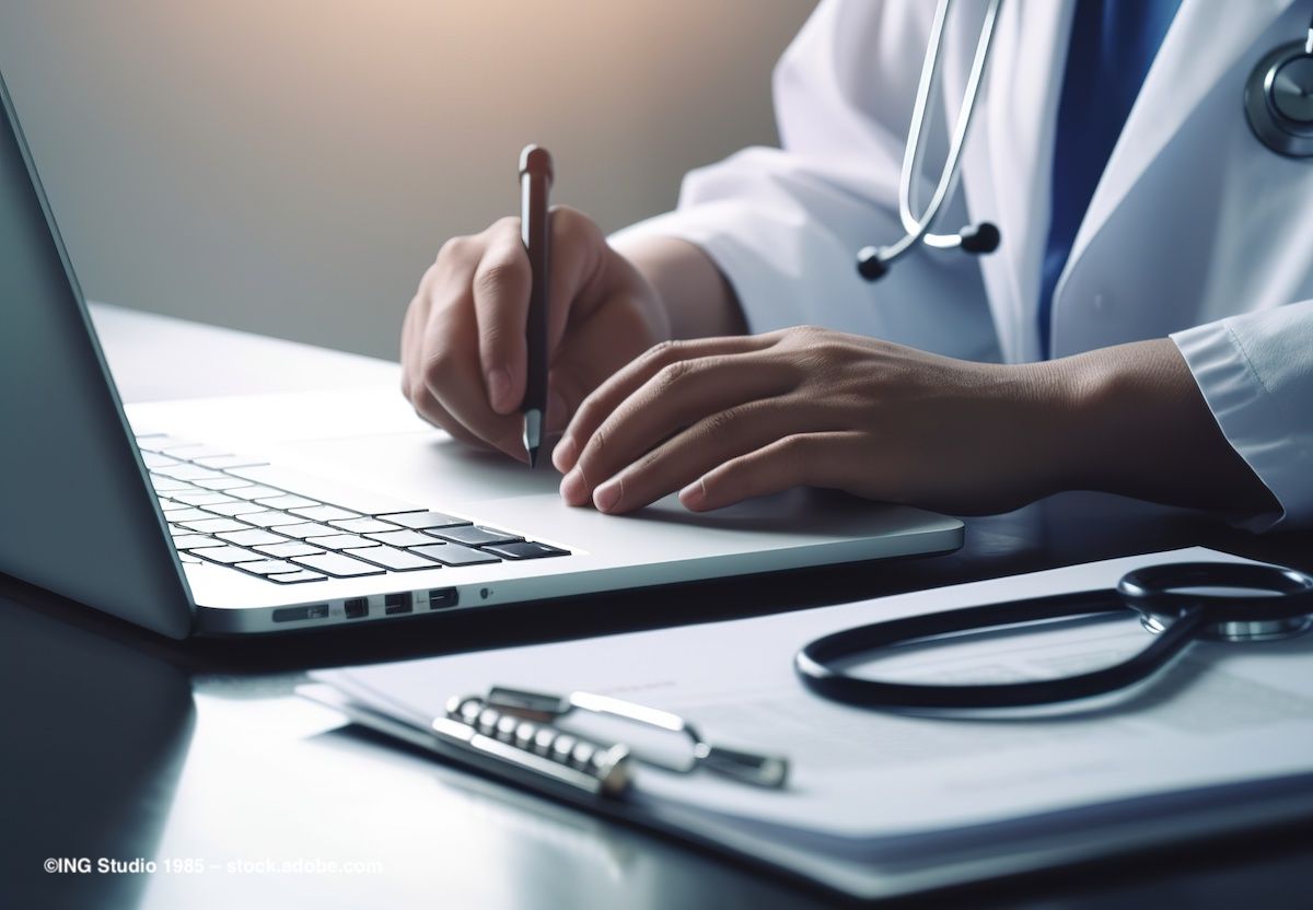 A doctor sits at a desk, writing on a clipboard and typing on a computer. Image credit: ©ING Studio 1985 – stock.adobe.com