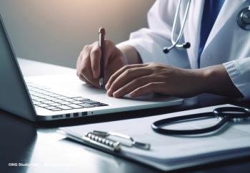 A doctor sits at a desk, writing on a clipboard and typing on a computer. Image credit: ©ING Studio 1985 – stock.adobe.com