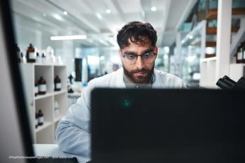 A scientist in a lab sits at a computer. He is looking at the screen of the computer, which is not visible to the camera. He wears glasses and a white lab coat. Image credit: ©PeakPoints/peopleimages.com – stock.adobe.com
