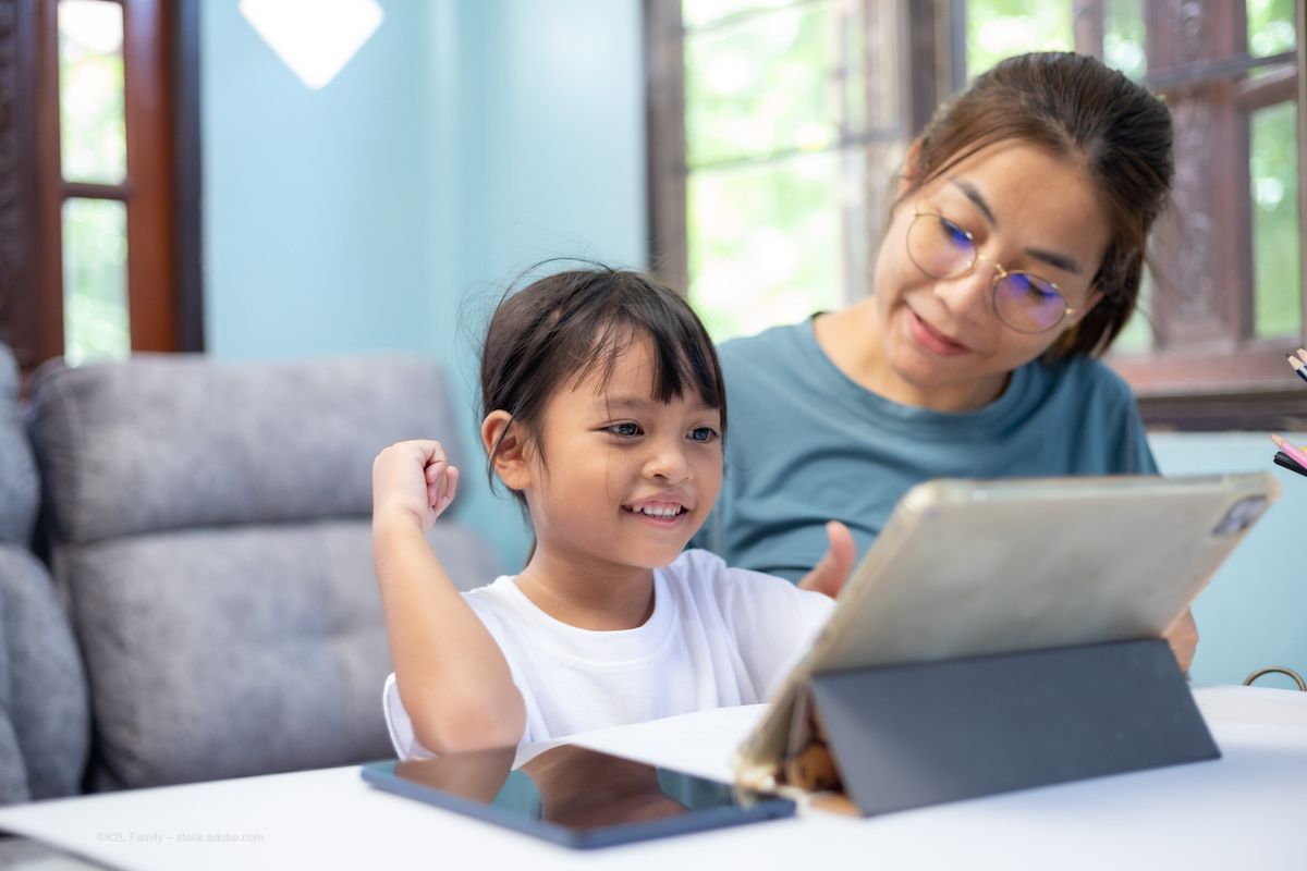 A woman with glasses helps a child, who may be her daughter, use a tablet. Concept image for online school, homeschool, screen time, myopia, and paediatric astigmatism. Image credit: ©K2L Family – stock.adobe.com