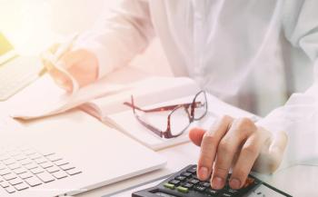 A person in a white lab coat sits with a computer and a calculator at hand.  Image credit: ©everythingpossible – stock.adobe.com