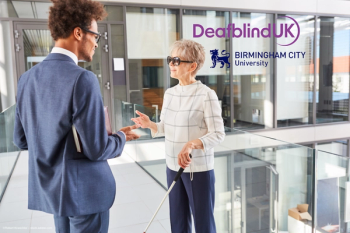 A pair of blind adults chat in an office building. The logos for Deafblind UK and Birmingham City University are emblazoned above their heads on the image. Image credit: ©Robert Kneschke – stock.adobe.com
