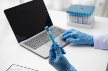 A person in a lab coat holds a test tube full of fluid. They enter data onto a laptop. Image credit: ©Parichat – stock.adobe.com